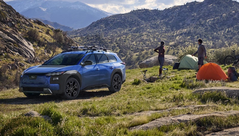 A static image of a blue 2026 Subaru Trailseeker parked on a grassy clearing in a rocky mountain landscape.