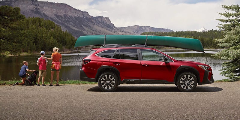 Red Subaru Outback parked beside a lake with a canoe secured on the roof and mountains in the background