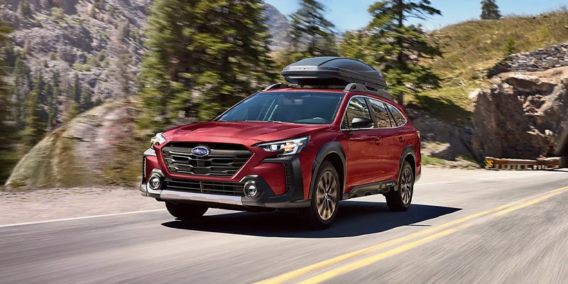 Red Subaru Outback traveling on a scenic mountain road with a roof-mounted cargo carrier
