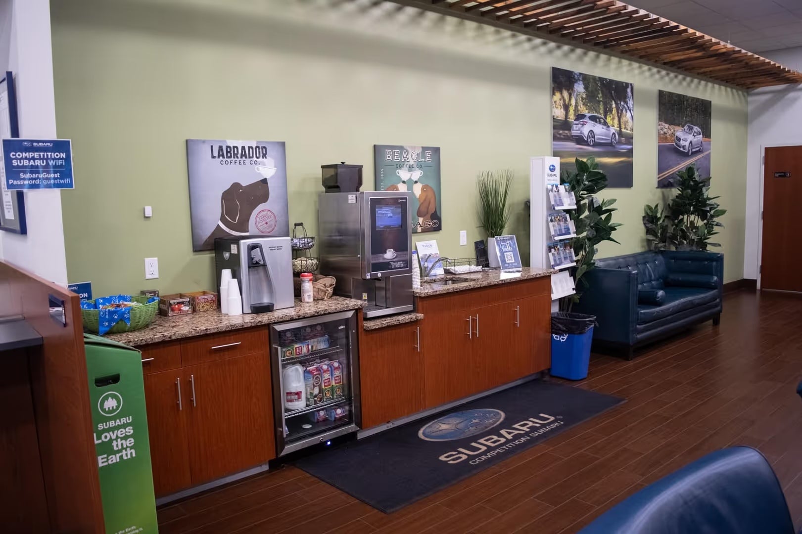 Subaru dealership waiting area with a self-serve coffee station, mini-fridge, dog-themed posters, and a blue couch.
