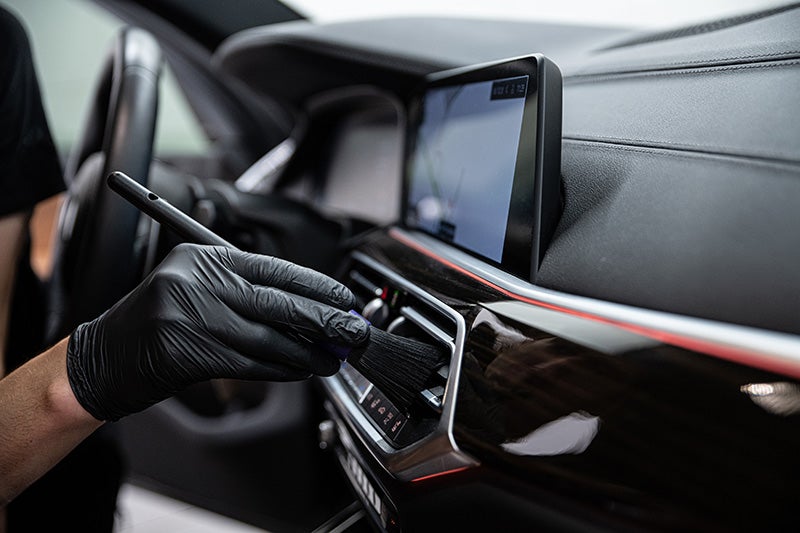 Close-up of a gloved hand using a detailing brush to clean the dashboard and air vent area inside a car