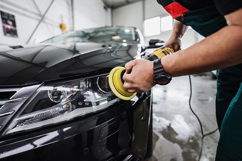 A technician is using an electric polishing machine with a yellow polishing pad on the front headlight area
