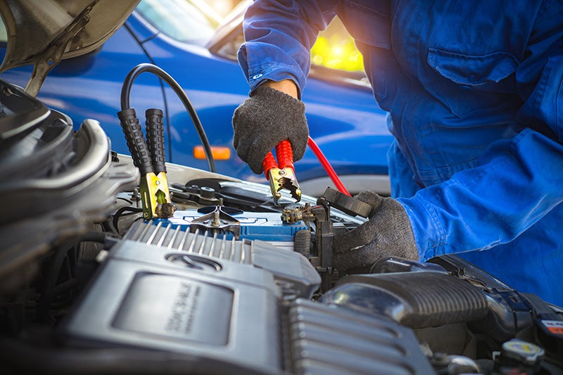 Technician connecting jumper cables to a vehicle battery inside an open engine compartment