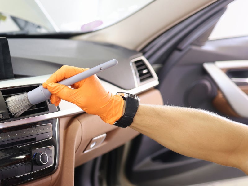 A hand in an orange glove uses a soft brush to clean a car's dashboard air vent