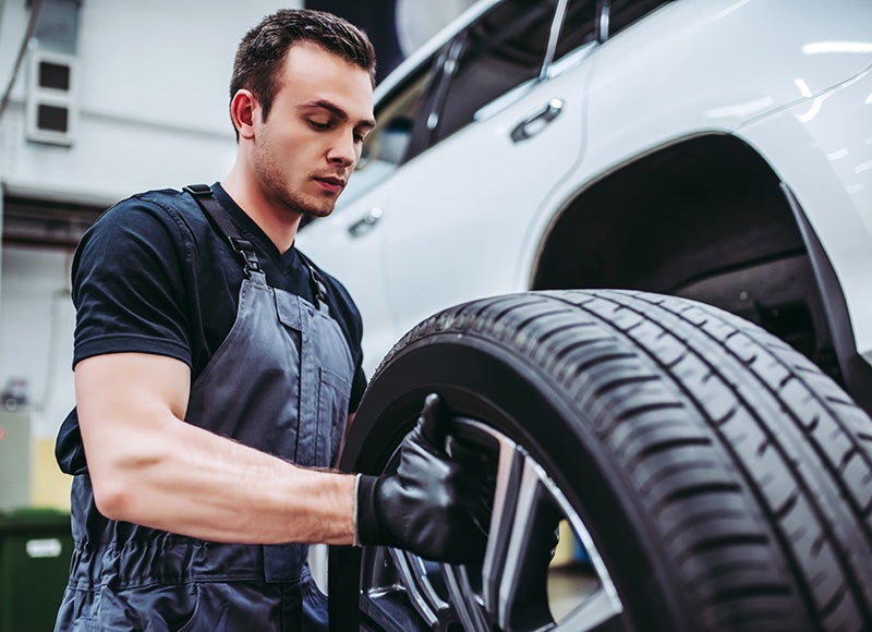 Automotive technician installing or inspecting a tire beside a vehicle in a service bay.